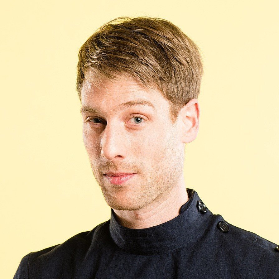 Portrait of a young man with short light brown hair and blue eyes against a yellow background.