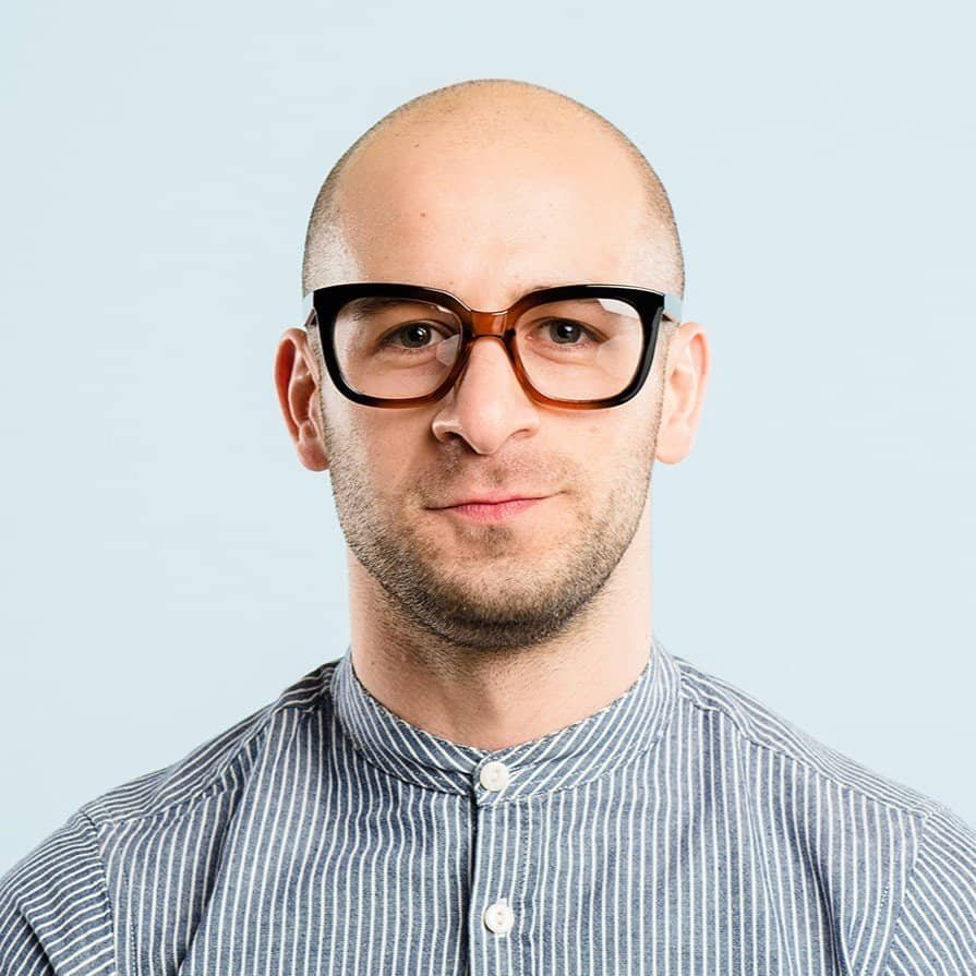 Portrait of a young man with glasses and a striped shirt against a light background.